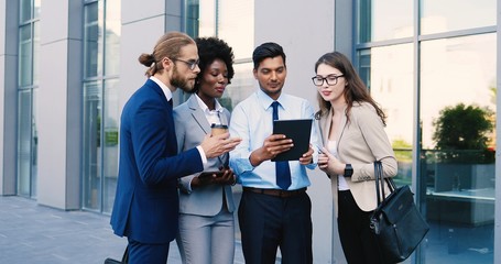 Multi ethnic team of young males and females white-collars workers standing outdoors at business center and watching tablet device. Mixed-races men and women with coffee and gadget at street.