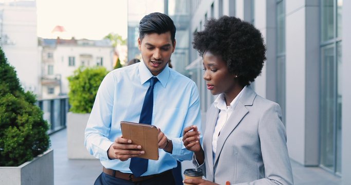 Hindu businessman showing something on tablet device to African American businesswoman with coffee at street in the morning, talking and discussing work. Mixed-races couple of business partners.