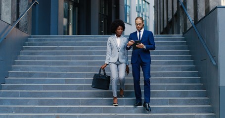Caucasian young male assistant showing something on tablet device to female African American boss. Mixed-races business partners walking down steps and talking. Using gadget. Leaving office building.
