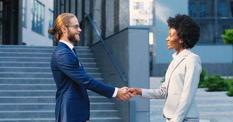 Caucasian young businessman and African American businesswoman shaking hands outdoors when meeting. Business mixed-races male and female partners at city street. Multi ethnic man and woman making deal