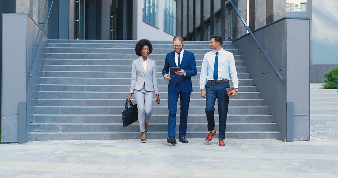 Cheerful multi ethnic young men and woman leaving business office building and stepping down the steps. Outdoors. African American businesswoman and mixed-races businessmen talking and using tablet.