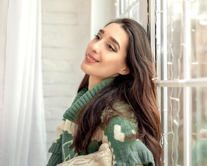 Portrait of young pretty female sitting on windows with Christmas garland and white curtains