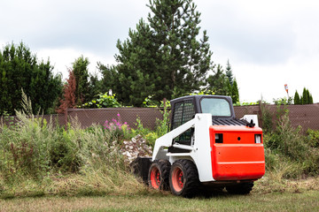 Naklejka premium A skid steer loader clears the site for construction. Land work by the territory improvement. Machine for work in confined areas. Small tractor with a bucket for moving soil and bulk materials.
