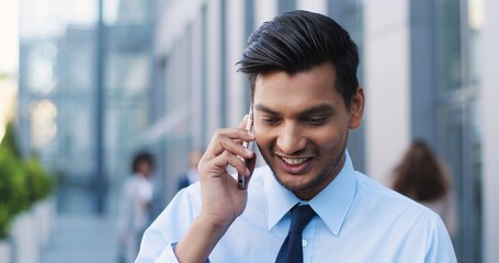 Hindu cheerful young businessman in tie standing at street and talking on phone. Man in business style speaking on mobile phone and laughing. Smiled happy male white-collar employee. Close up.