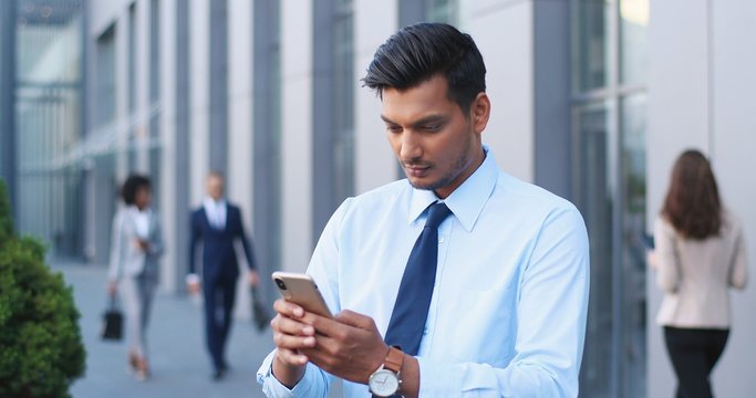 Handsome Hindu Young Businessman In Tie Using Smartphone And Texting Message At Street. Man From India In Business Style Tapping And Scrolling On Mobile Phone. Outdoors. Close Up.