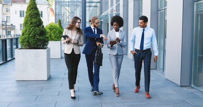 Mixed-races young cheerful people in business style walking the street , laughing and talking. Multiethnic business white-collars males and females smiling and going to work in morning. Men and women.