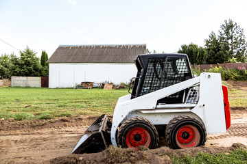 A skid steer loader clears the site for construction. Land work by the territory improvement. Machine for work in confined areas. Small tractor with a bucket for moving soil and bulk materials.