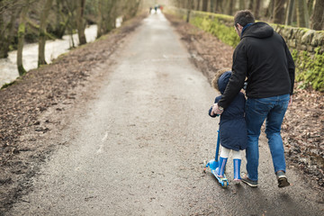 A father helps her daughter ride her push scooter along a footpath near a river with trees before sunset in the Pentland hills regional park in Edinburgh, Scotland, United Kingdom