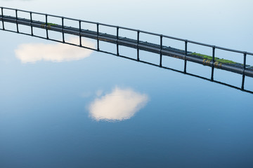 Fototapeta na wymiar A blue sky with clouds reflected on the calm water of a reservoir in the Pentland Hills Regional Park, Edinburgh, Scotland, United Kingdom, with a metallic runway across the calm water