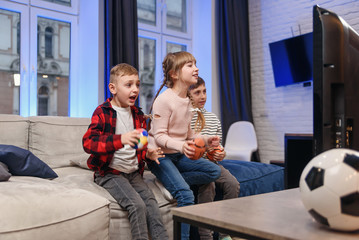 Funny three child friends are sitting on the couch at home and enjoying soccer match and shouting when see scored ball.