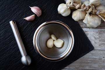 Garlic cloves in a metal mortar ready to be crushed with a pastel on black stone background placed on wooden table with braided garlic on top right