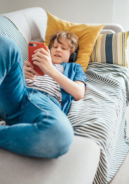 Preteen boy lying at home on cozy sofa dressed casual jeans and new sneakers listening to music and chatting using wireless headphones connected with smartphone. Child use electronic devices concept