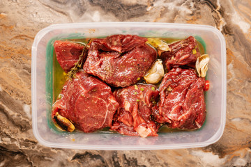 Marinated beef steak with butter, garlic and rosemary in a plastic food container on a marble background.