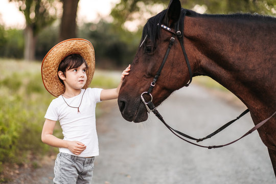 Cute little boy in a cowboy hat stroking a beautiful horse outdoors