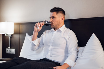 A handsome young businessman drinking red wine in bed.