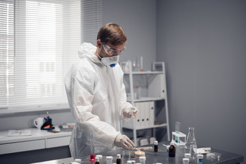 A scientist in protective eyewear, respirator and white overalls standing at the table in the science laboratory, working on something.