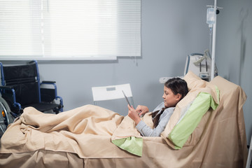 Little girl with an ice cream and a tablet in a hospital bed, her wheelchair is beside her bed.
