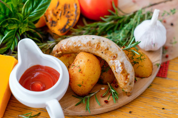 Delicious sausage grilled with potatoes and yellow tomatoes, ketchup is standing in a white cup in front.