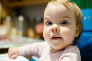 Photo portrait of a baby girl with pink cheeks she sits in a chair for feeding and eats porridge. Child sitting at home