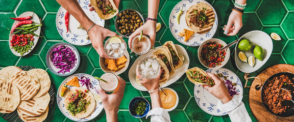 Friends home taco party. Flat-lay of Mexican traditional dishes Tacos with beef meat, corn tortillas , tomato salsa and peoples hands with glasses over green background, top view. Mexican cuisine - obrazy, fototapety, plakaty