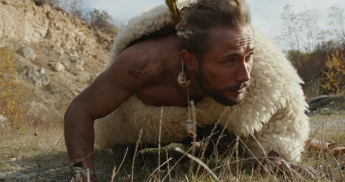 Low angle of bearded man in animal hide licking and smelling soil while stalking prey against cloudy sky in countryside