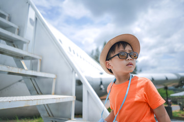 portrait of cute small Asian boy wearing hat and sunglasses standing next to airplane during family vacation in summer time 