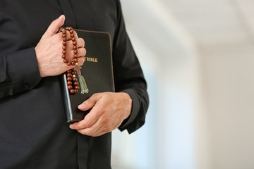 Male priest with Bible and rosary beads at home, closeup