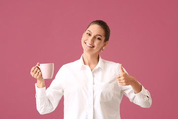 Beautiful young woman with cup of tea showing thumb-up on color background