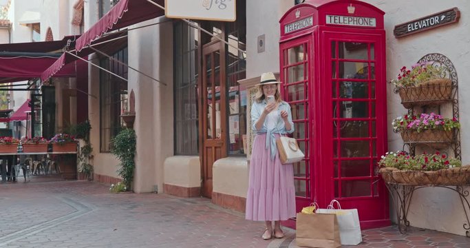 Cheerful smiling woman is having fun and making selfie on smartphone at red London telephone booth on a street of old town in European country. Stylish traveler is taking picture with London landmark