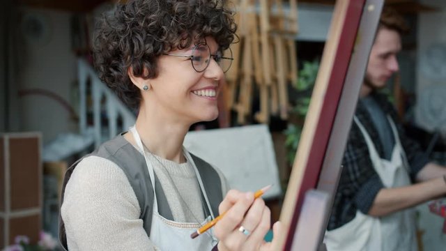 Happy young woman is drawing picture on easel smiling enjoying creative lesson in art studio while teacher is teaching serious young man in background
