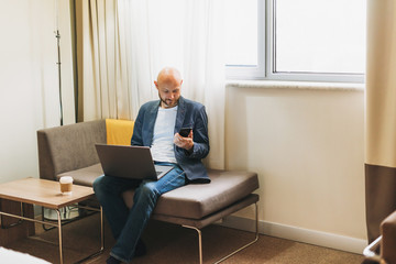 Adult bald bearded man in blue jacket working on laptop in hotel room