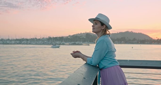 Traveler in trendy casual outfit enjoying cinematic pink sunset on ocean pier. Slow motion of relaxed woman contemplating scenic water view during vacation. Female traveler on summer trip, 4K USA