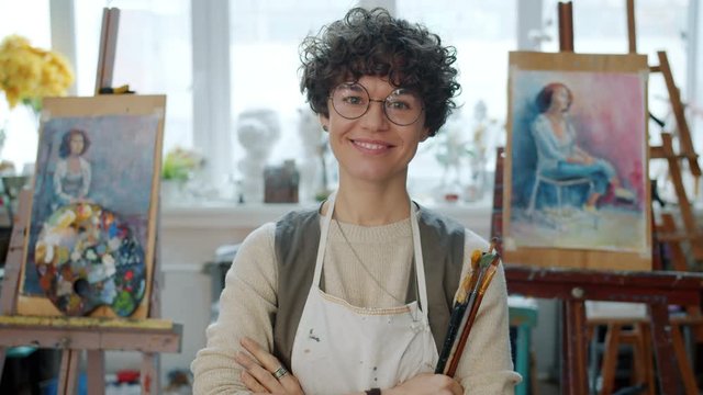 Portrait of young female painter standing in modern painting studio holding brushes looking at camera smiling wearing casual clothing and apron