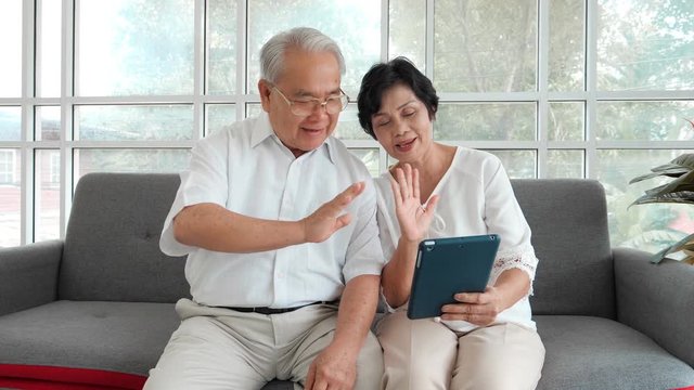 Pretty happy smiling Asian senior elderly couple talking on video on tablet while sitting on sofa in room. Old couple of joyful waving to camera while videochatting on device. Home concept