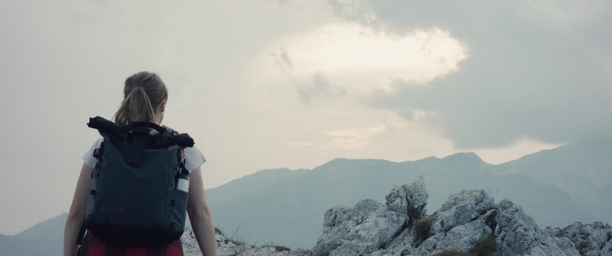 Active woman on a hike walk in the mountains hiking in fall or autumn cloudy evening climb up to the top rock lift extend arms at the view slowmotion light through cloud silhouette