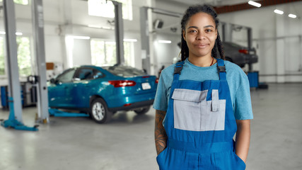 Add new life to your car. Portrait of young african american woman, professional female mechanic in uniform smiling at camera, standing in auto repair shop. Car service, repair, maintenance concept