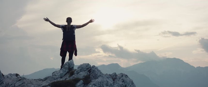 Woman on a hike in the mountains hiking in fall or autumn cloudy evening at the top rock lift extend arms at the view slowmotion light through clouds silhouette