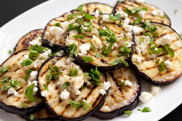 Homemade Grilled Eggplant with Feta and Herbs on a white plate on a black surface, side view. Close-up.