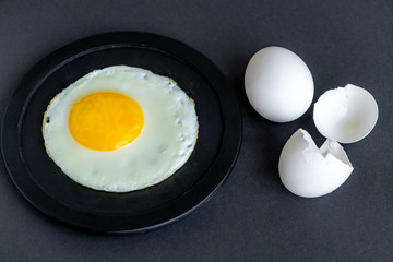 Fried egg. Close-up of fried eggs on a black plate. A whole egg and shells nearby, on a black background. Soft focus.
