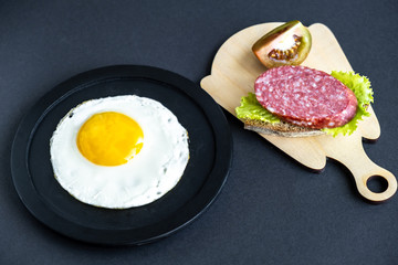 Scrambled eggs on a black plate on a black background, a sandwich with sausage, herbs and a slice of tomato. Soft focus.