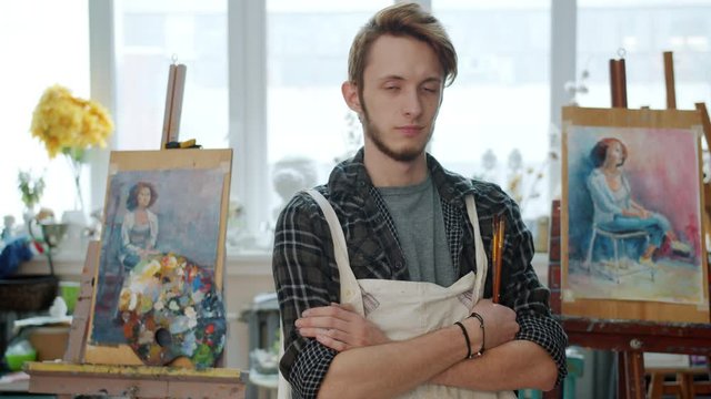 Portrait of creative young man artist standing in painting studio holding brushes looking at camera with serious face. Youth culture and hobby concept.