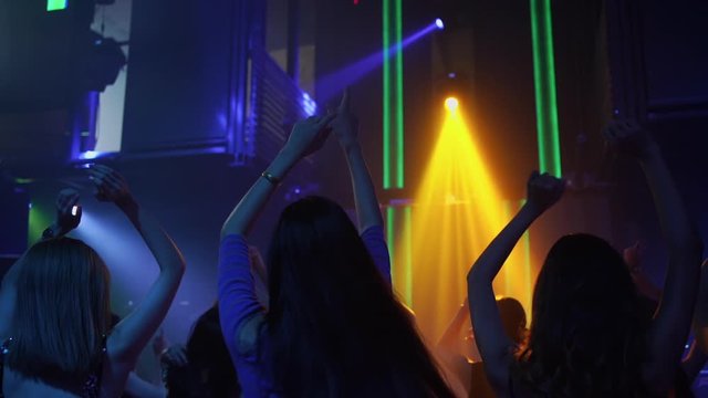 Rear view group of young beautiful woman dancing to the music played by dj with multi-color illuminated at night club. People enjoy nightlife and having fun with dance party at disco nightclub.
