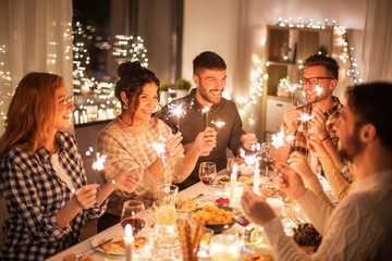 holidays, celebration and people concept - happy friends with sparklers having christmas dinner at...