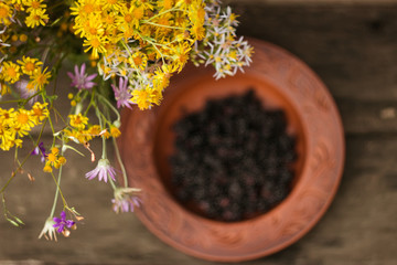 A round plate of red clay with blackberries stands on an old wooden table next to a bouquet of wildflowers; blurred background