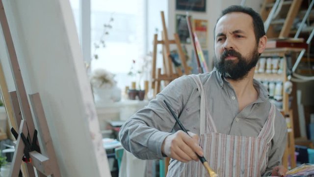 Slow motion of attractive bearded man painter working at picture painting indoors in studio concentrated on creative work. People and occupation concept.