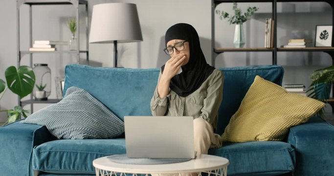 Arabian young woman in black hijab and glasses videochatting on laptop computer and sneezing or caughing in napkin on couch in living room. Sick female in headscarf talking on webcam and feeling bad.