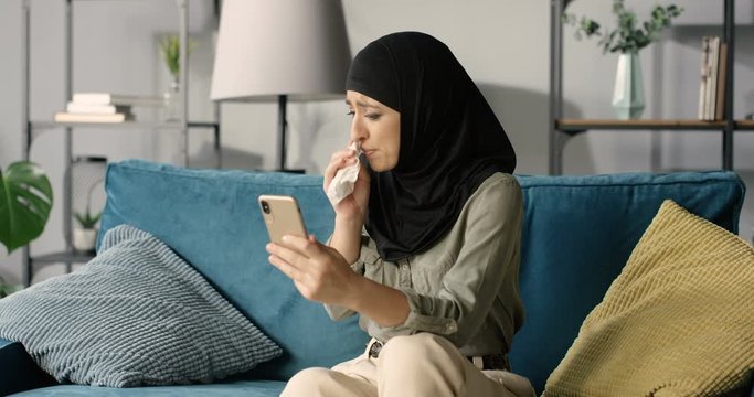 Arabian young woman in black hijab videochatting on smartphone and sneezing or caughing in napkin on couch in living room. Sick muslim female in headscarf talking on phone webcam and feeling bad.