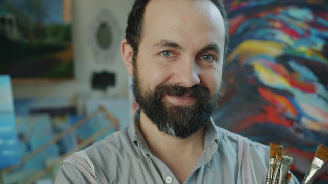 Close-up portrait of attractive bearded man artist standing in colorful studio with pictures holding brushes looking at camera with happy smile