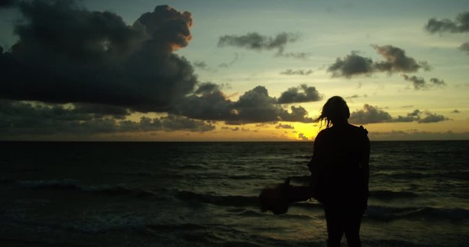 Woman stands against a backdrop of rolling clouds and an incredible sunset looking out at ocean as her dress billows in the wind on the edge of cliff