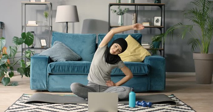 Arabian woman in hijab sitting on yoga mat on floor in front of the laptop computer at home. Attractive slim muslim girl working out indoor with internet video course. Lesson with instructor online.